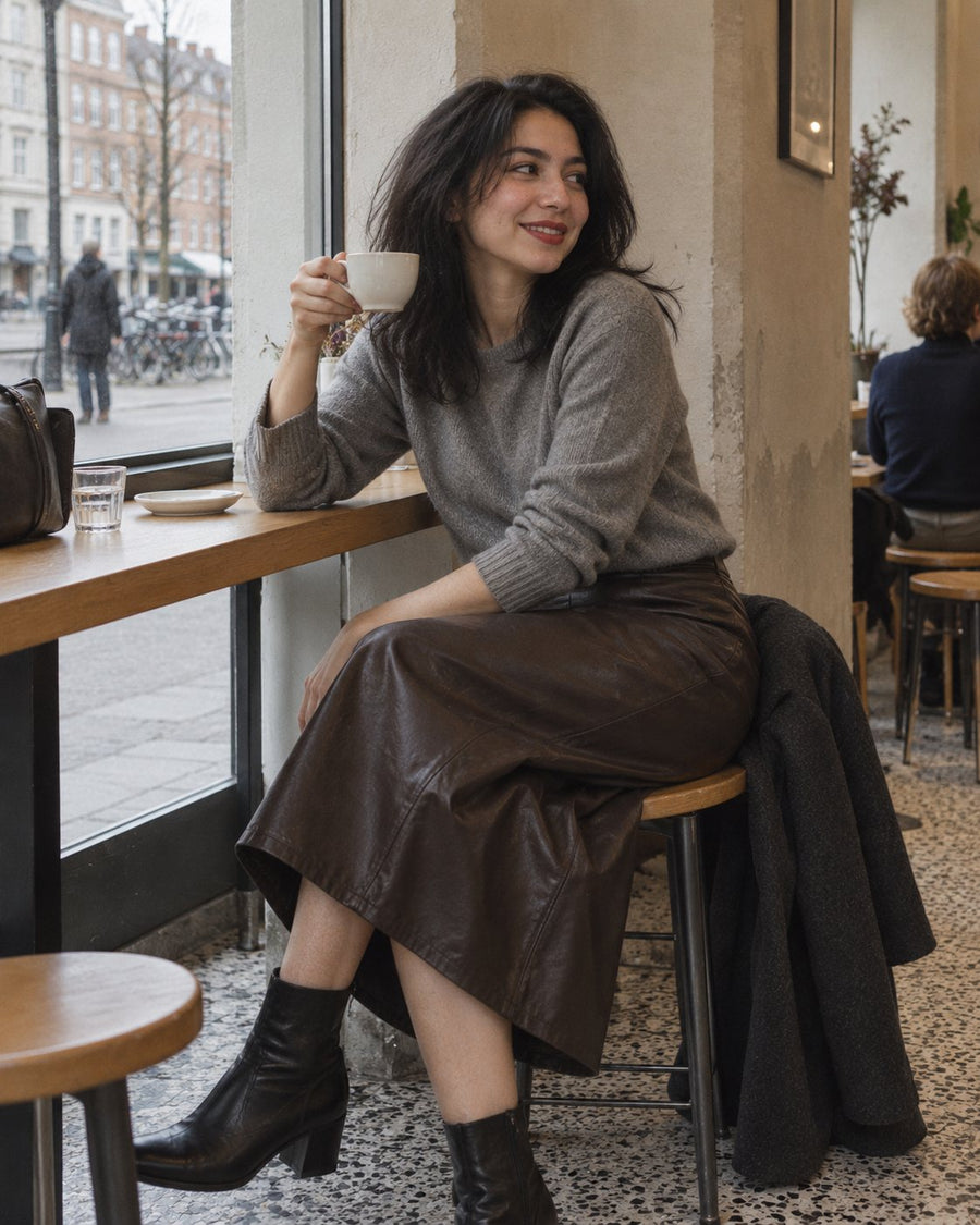 Woman in a dark brown leather midi skirt holding a cup at a café window counter